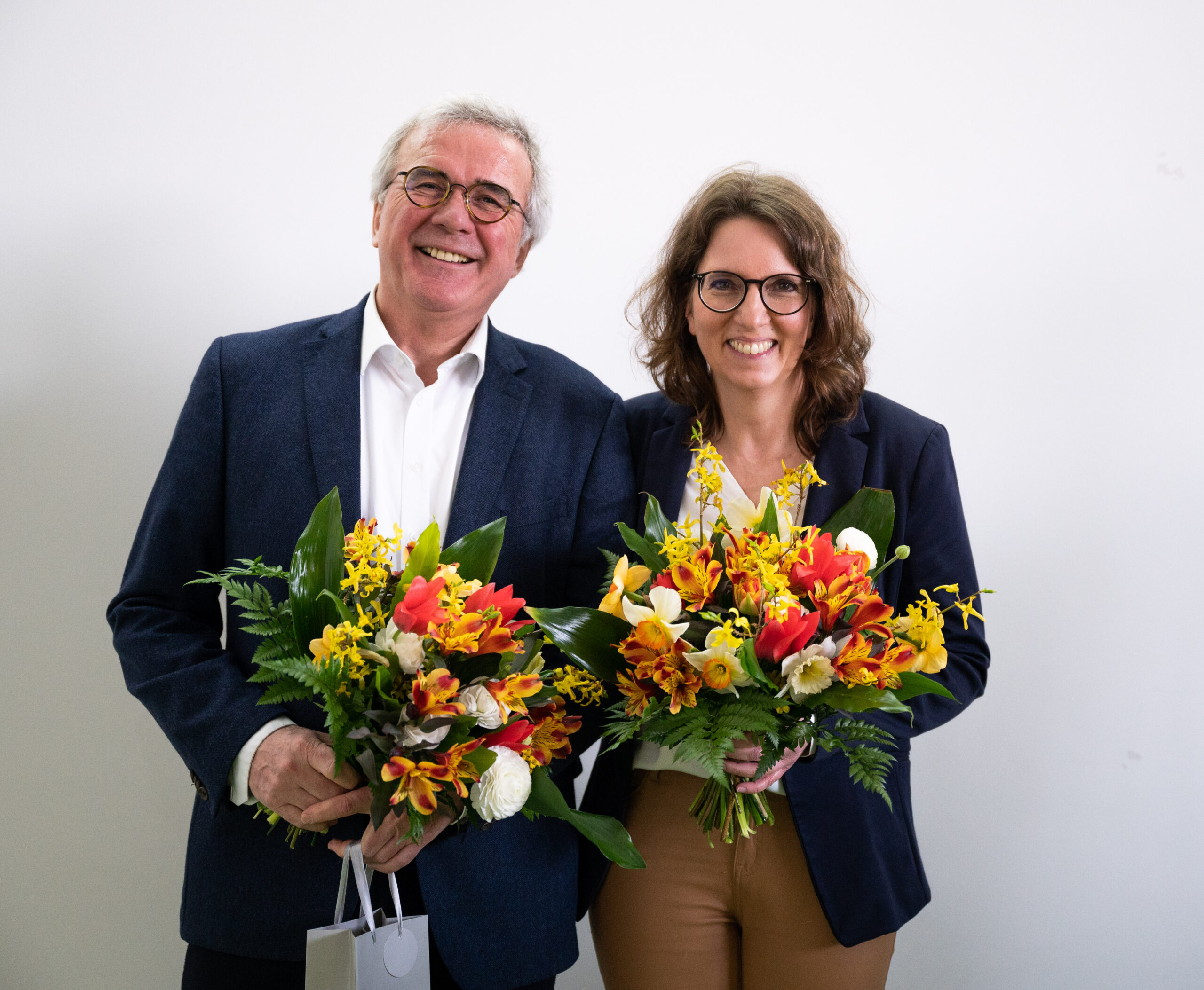 Ein Mann und eine Frau, beide in Business-Kleidung und mit Brille, lächeln und halten bunte Blumensträuße in den Händen. Sie stehen vor einem schlichten weißen Hintergrund und verkörpern den lebendigen Geist der Hochschule Coburg.