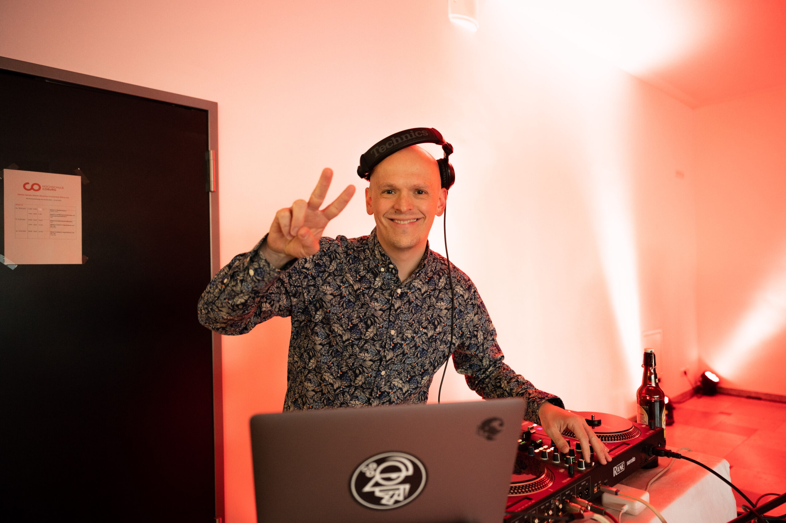 Ein DJ mit Kopfhörern, der die Hochschule Coburg vertritt, lächelt und hält ein Peace-Zeichen hoch, während er hinter einem Laptop und DJ-Equipment steht. Der Raum ist sanft in warmen Farben beleuchtet und auf dem Tisch daneben steht eine Flasche.