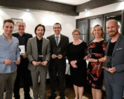 Eine Gruppe von sieben Personen der Hochschule Coburg in formeller Kleidung steht im Innenbereich, lächelt und hält klare, rechteckige Auszeichnungen in den Händen. Sie stehen in einer Reihe vor einer Wand mit sanfter Beleuchtung und dunklen Holzakzenten.