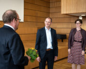 Drei Personen befinden sich in einem Besprechungsraum der Hochschule Coburg. Ein Mann im Vordergrund hält einen kleinen Blumenstrauß in der Hand. Zwei weitere Personen, ein Mann und eine Frau, stehen ihm lächelnd in Geschäftskleidung gegenüber. Die Einrichtung des Raums besteht aus eleganten Holzwänden und -vertäfelungen, die zur formellen Atmosphäre beitragen.
