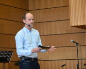 Ein Mann im hellblauen Hemd steht in einem holzgetäfelten Raum der Hochschule Coburg, spricht in ein Headset-Mikrofon und gestikuliert mit den Händen. In der Hand hält er ein Notizbuch, im Hintergrund sind ein Mikrofonständer und ein Notenpult zu sehen.