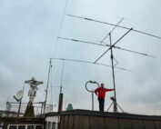 Eine Person steht auf einem Dach der Hochschule Coburg neben einer großen Radioantenne unter einem bewölkten Himmel. In der Nähe sind verschiedene Satellitenschüsseln und -geräte verstreut, in der Ferne bilden Gebäude und Bäume den Hintergrund.