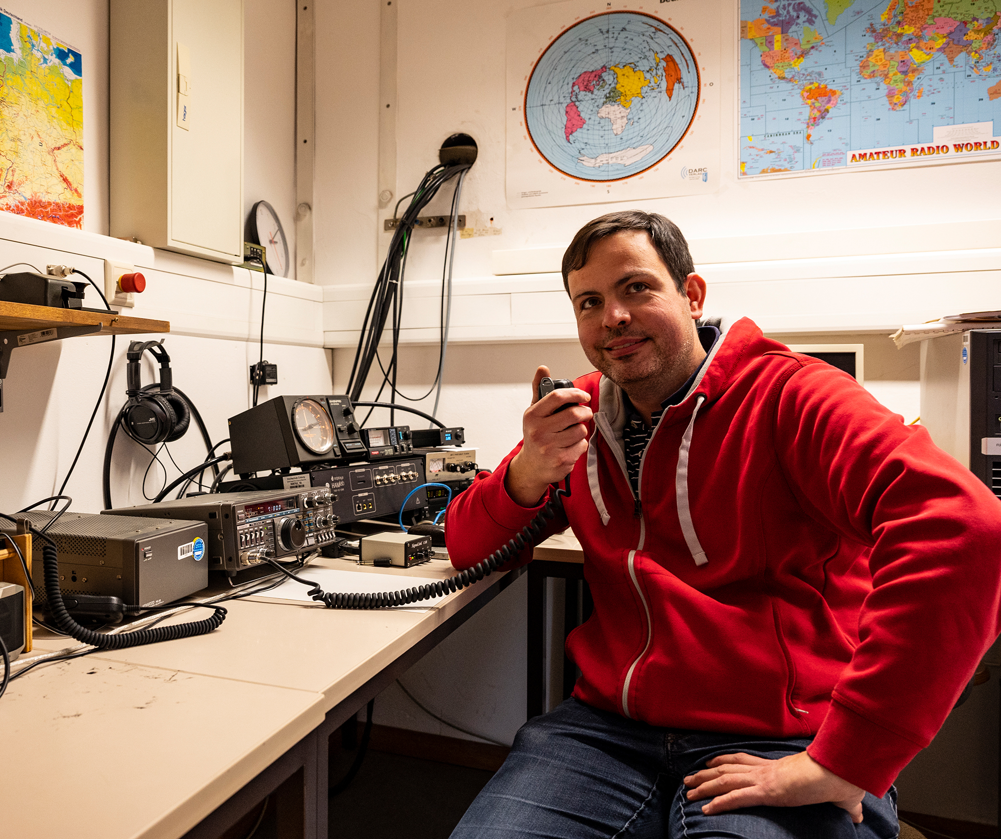 Ein Mann in einem roten Kapuzenpulli sitzt am Pult eines Radiosenders, ist mit einer Funkmaschine ausgestattet und kommuniziert, während Weltkarten die Wände schmücken. Im Rahmen seines Projekts an der Hochschule Coburg vertieft er sich in die Kunst des Rundfunks und verfeinert seine Fähigkeiten mit jedem Wort, das er ins Mikrofon spricht.
