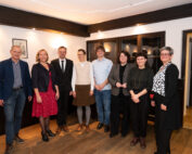 Eine Gruppe von acht Personen, vermutlich von der Hochschule Coburg, steht in einer Reihe im Innenbereich. Sie tragen halbformelle bis legere Kleidung. Sie lächeln herzlich in einem Raum mit Holzboden und einem gerahmten Bild an der Wand.