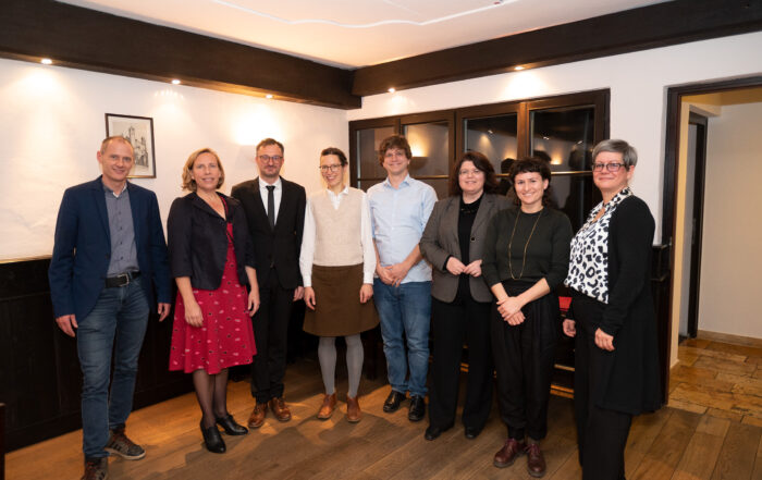 Eine Gruppe von acht Personen, vermutlich von der Hochschule Coburg, steht in einer Reihe im Innenbereich. Sie tragen halbformelle bis legere Kleidung. Sie lächeln herzlich in einem Raum mit Holzboden und einem gerahmten Bild an der Wand.