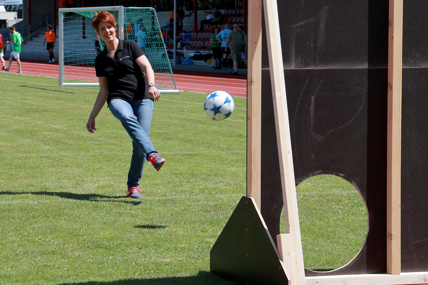 Eine Person in schwarzem Hemd und Jeans kickt einen Fußball in Richtung einer Holzplatte mit einem kreisrunden Loch. Die Szene spielt auf der Wiese der Hochschule Coburg und zeigt ein Fußballtor und Menschen im Hintergrund.