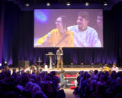 Ein Redner steht auf einer Bühne der Hochschule Coburg vor einem großen Publikum. Auf einer großen Leinwand ist eine Nahaufnahme einer jungen Frau in einem gelben Kapuzenpullover zu sehen, die ein Mikrofon hält, neben ihr ein Mann. Violettes Licht erfüllt den Raum.