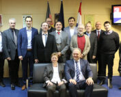 Eine Gruppe von dreizehn Personen in Business-Kleidung posiert für ein Foto an der Hochschule Coburg. Zwei Personen sitzen vorne, die anderen stehen dahinter. Sie befinden sich in einem Raum mit gerahmten Zertifikaten und einem digitalen Schild an der Wand.