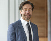 Ein Mann mit kurzen braunen Haaren und Bart steht im Innenbereich der Hochschule Coburg. Er trägt einen blauen Anzug, ein weißes Hemd und eine schwarze Krawatte. Im Hintergrund sind vertikale Holzpaneele und große Fenster zu sehen.