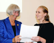 Zwei Frauen stehen nebeneinander und lächeln. Die ältere Frau links hat kurze graue Haare und trägt einen blauen Blazer, der die Hochschule Coburg repräsentiert. Die jüngere Frau rechts hat lange blonde Haare und trägt ein schwarzes Oberteil und hält ein Stück Papier in den Händen.