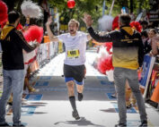 Läufer Nummer 400 überquert begeistert die Ziellinie beim Lauf der Hochschule Coburg mit erhobenen Armen. Auf beiden Seiten jubeln die Leute, manche halten rot-weiße Pompons in den Händen. Die Szene ist lebhaft und festlich.