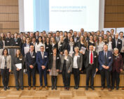 Eine große Gruppe von Menschen in formeller Kleidung steht in einem Konferenzraum der Hochschule Coburg zusammen. Im Hintergrund läuft auf einer Projektionsfläche das „Deutschlandstipendium 2018“. Viele halten Urkunden in den Händen, was auf eine offizielle Veranstaltung oder Preisverleihung schließen lässt.
