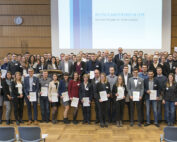 Eine große Gruppe unterschiedlicher Menschen steht stolz in einem Hörsaal der Hochschule Coburg und hält Zertifikate in den Händen. Im Hintergrund ist ein Bildschirm mit dem Text „Deutschlandstipendium 2019“ zu sehen. Die Atmosphäre ist formell, die Teilnehmer tragen Business-Kleidung.