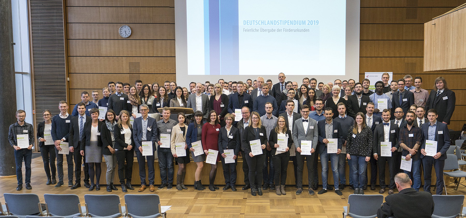 Eine große Gruppe unterschiedlicher Menschen steht stolz in einem Hörsaal der Hochschule Coburg und hält Zertifikate in den Händen. Im Hintergrund ist ein Bildschirm mit dem Text „Deutschlandstipendium 2019“ zu sehen. Die Atmosphäre ist formell, die Teilnehmer tragen Business-Kleidung.