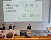 In einem modernen Hörsaal der Hochschule Coburg findet ein Vortrag statt, auf einer großen Leinwand werden Informationen zur Diensttagsreihe Coburg und ihrer 20-jährigen Jubiläumsveranstaltung am 27.05.22 um 13:00 Uhr angezeigt. Das Publikum sitzt und hört aufmerksam zu.