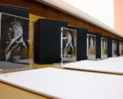 Auf einem Tisch in der Hochschule Coburg ist eine Reihe gläserner Auszeichnungen mit eingravierten Profilen ausgestellt. Jede Auszeichnung steht vor einer schwarzen Box, darunter liegen weiße Umschläge. Im Hintergrund befinden sich eine Holzwand und eine Projektionsfläche.