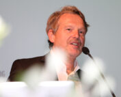 Ein Mann im Anzug spricht in ein Mikrofon an der Hochschule Coburg. Er hat kurzes, helles Haar und lächelt leicht. Der Hintergrund ist schlicht und größtenteils verschwommen, teilweise ist ein undeutlicher Text zu sehen.