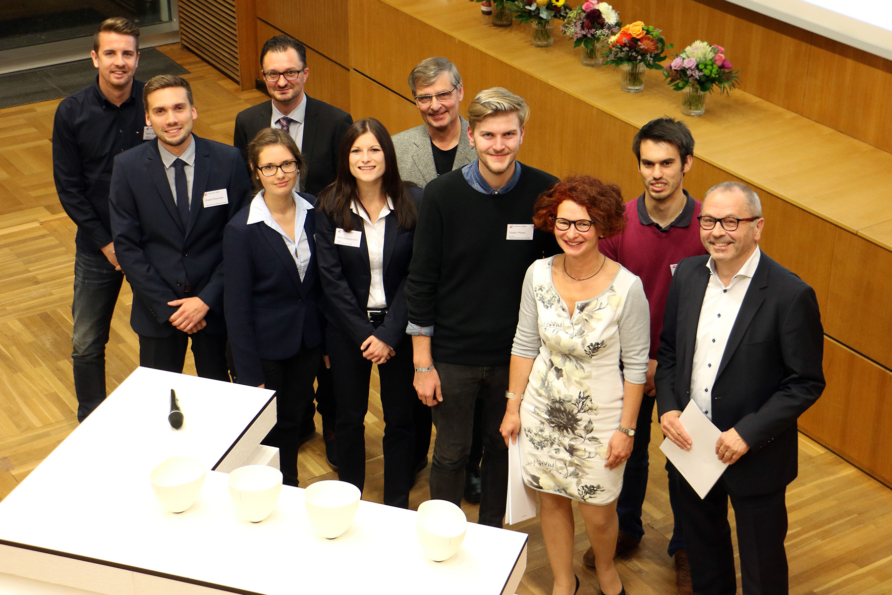 Eine Gruppe von Fachleuten, darunter Männer und Frauen, posiert für ein Gruppenfoto in einem formellen Rahmen an der Hochschule Coburg. Sie stehen auf einem Holzboden neben einem Tisch mit weißen Schüsseln, während ein Blumenarrangement den Hintergrund wunderschön schmückt.