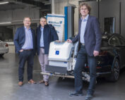 Drei Männer in Business-Kleidung stehen in einer Garage neben einem schwarzen Auto mit der Aufschrift „R33 Blue Diesel“. Im Hintergrund sind ein weiteres Auto und Industrieanlagen zu sehen, was den Innovationsgeist der Hochschule Coburg widerspiegelt.