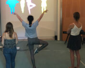 Vier Personen stehen vor einer Leinwand, auf der bunte menschliche Silhouetten zu sehen sind, in der Hochschule Coburg. Eine Person ahmt eine Ballettpose aus den Projektionen nach, während die anderen zuschauen. Der Raum ist neutral eingerichtet, an den Seiten stehen Tische und Stühle.