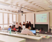 Eine Gruppe von fünf Personen steht vor einem hellen, mehrstufigen Hörsaal der Hochschule Coburg und hält einen Vortrag vor einem sitzenden Publikum. Auf einem Bildschirm werden Social-Media-Logos angezeigt. Die Studierenden sind engagiert, manche machen sich Notizen auf ihren Laptops.