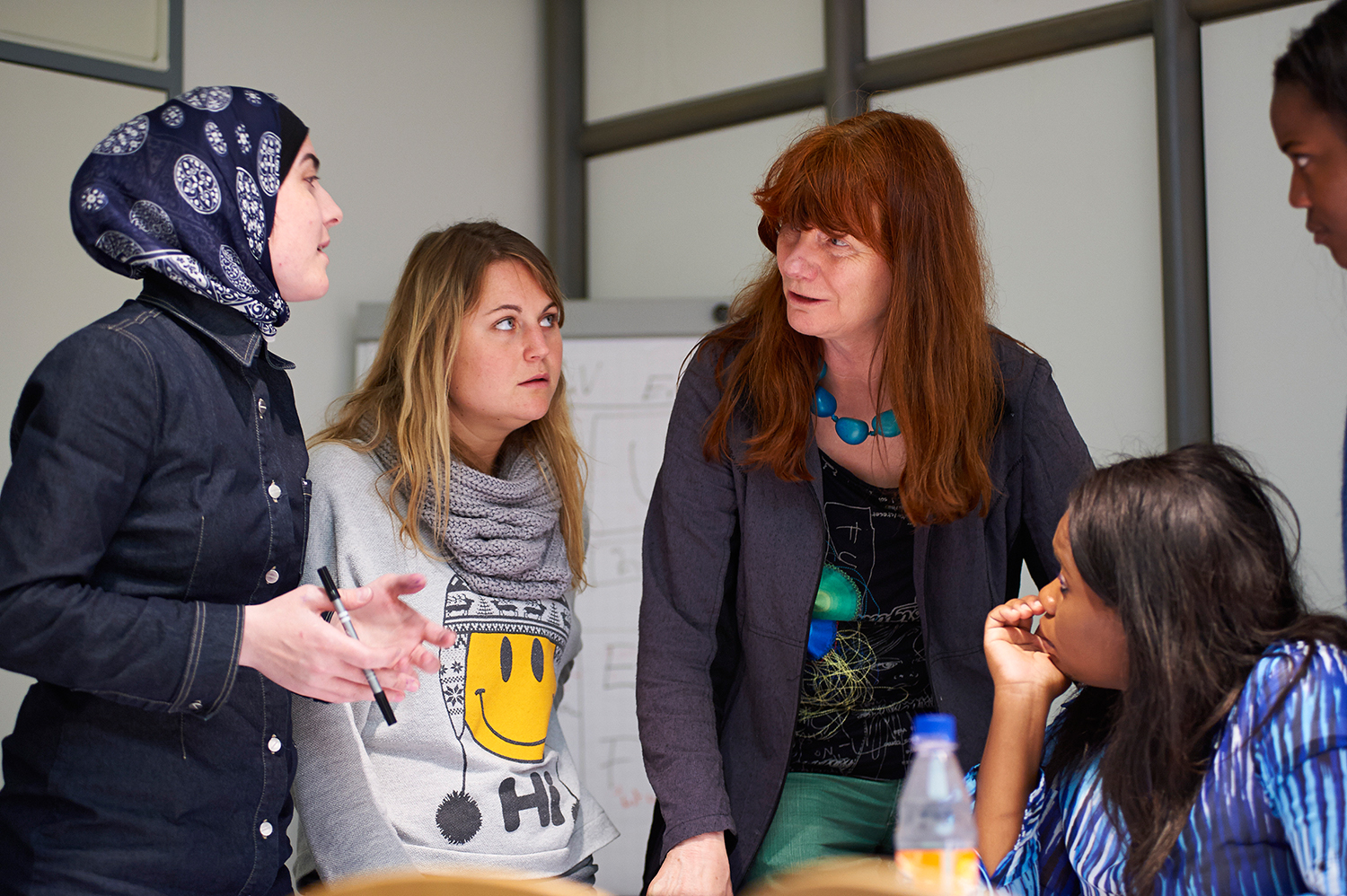 Im Büro der Hochschule Coburg diskutieren vier Personen angeregt. Eine trägt Hijab, eine andere hat lange rote Haare und eine Halskette, die dritte hat hellbraune Haare und ein Kopftuch. Die vierte sitzt und hört aufmerksam den unterschiedlichen Perspektiven zu.