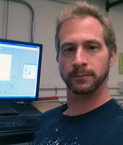 Eine Person mit kurzen Haaren und Bart, möglicherweise ein Student der Hochschule Coburg, sitzt vor einem Computer, auf dem ein technisches Diagramm angezeigt wird. Die Umgebung scheint ein Büro oder Labor zu sein. Sie trägt ein dunkles T-Shirt und blickt selbstbewusst in die Kamera.