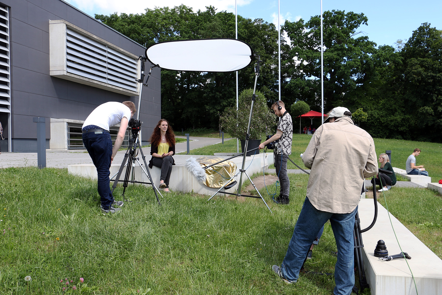 Eine Frau wird im Freien auf einer Wiese in der Nähe des modernen Gebäudes der Hochschule Coburg interviewt. Ein Team filmt mit Kamera, Mikrofon und Beleuchtungsausrüstung. Im Hintergrund sind einige Menschen zu sehen, die zwischen Bäumen unter einem weiten Himmel sitzen und gehen.