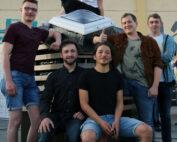 Eine Gruppe von sechs Personen, fünf Männer und eine Frau, posiert gemeinsam im Freien vor einem Solarpanel der Hochschule Coburg. Einige sitzen, andere stehen und alle lächeln in die Kamera. Sie sind lässig in Jeans und T-Shirts gekleidet und spiegeln die entspannte Atmosphäre auf dem Campus wider.