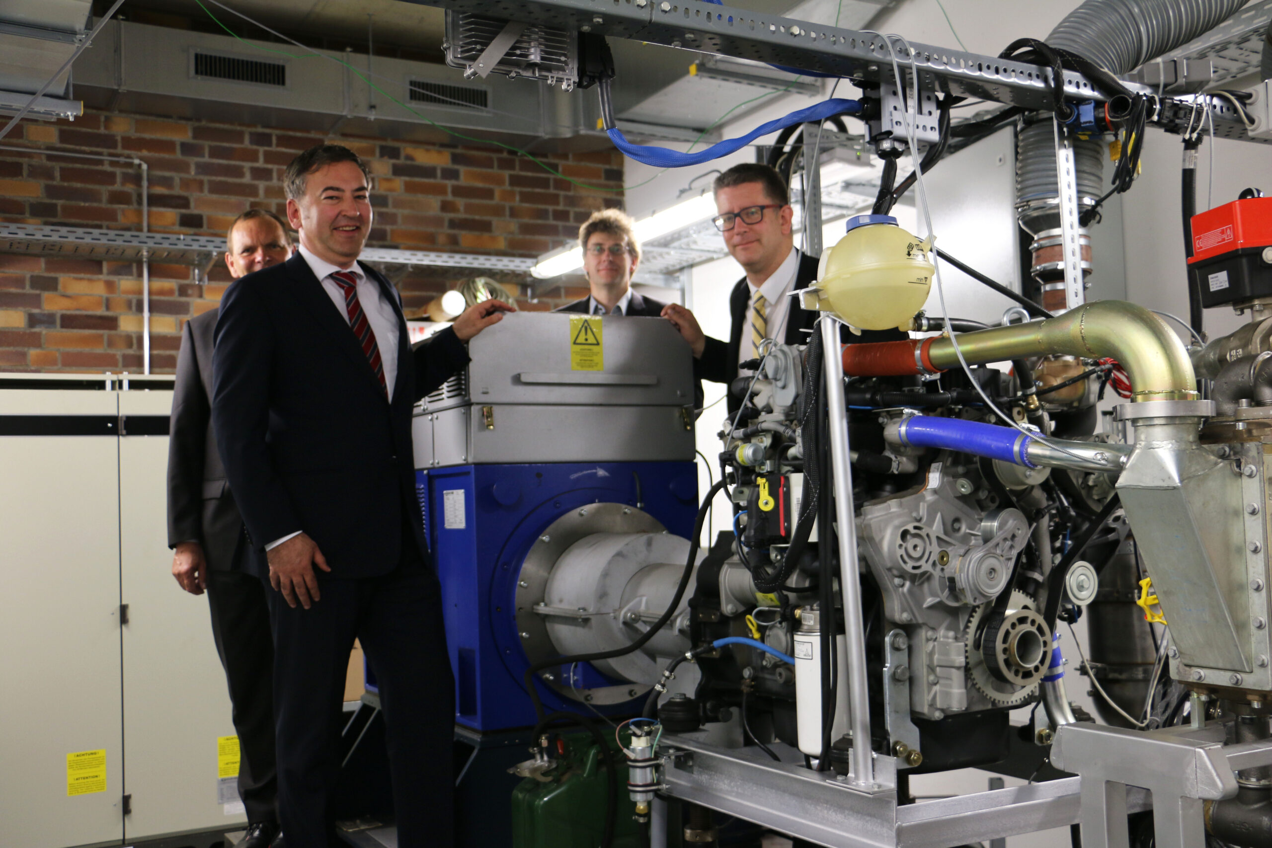 Vier Männer in Anzügen stehen lächelnd um Industriemaschinen in einer Fabrik herum. Im Hintergrund sind Rohre und elektrische Geräte der Hochschule Coburg zu sehen. Ein Mann deutet auf die Maschinen und deutet damit eine Präsentation oder Erklärung an.
