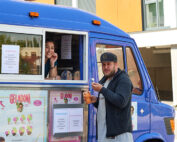Eine lächelnde Frau serviert einem Mann mit Mütze durch das Fenster eines farbenfrohen Trucks mit Hochschulmotiven der Coburg ein Eis. Der blau-violette Truck ist mit Eisplakaten und Speisekarten geschmückt. Der Mann hält eine Waffel mit einer kleinen Kugel Eis in der Hand und genießt das aufregende Erlebnis.