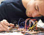 An der Hochschule Coburg konzentriert sich eine Person intensiv auf die Montage und Prüfung einer komplexen Platine mit zahlreichen bunten Drähten und Komponenten. Ihre Hände gehen behutsam mit der Elektronik um und verkörpern damit das Engagement der Universität für Präzision und Innovation im Ingenieurwesen.