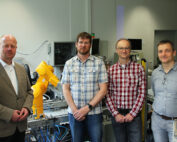 Vier Männer stehen in einem Labor der Hochschule Coburg, umgeben von Maschinen und einem gelben Roboterarm. Sie tragen eine Mischung aus Freizeit- und Businesskleidung und verkörpern damit die innovative Mischung aus Technologie und Ingenieurskunst, die das zukunftsorientierte Umfeld der Hochschule kennzeichnet.