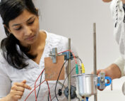 In einem Labor an der Hochschule Coburg arbeiten zwei Personen konzentriert an einem komplexen elektronischen Gerät mit Drähten und Komponenten. Einer deutet auf einen Teil des Geräts, während der andere es genau beobachtet. Beide sind konzentriert und arbeiten zusammen, was ihr Engagement für Innovation zeigt.