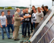 Eine Gruppe von Menschen steht auf einem Dach der Hochschule Coburg und hat sich um einen Mann im Anzug versammelt, der auf eine Reihe von Solarmodulen zeigt. Die Solarmodule sind deutlich im Vordergrund zu sehen und ragen in den Himmel.