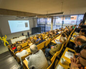 In einem vollen Hörsaal der Hochschule Coburg sitzen die Teilnehmer in Reihen aus Holzstühlen. Zwei Vortragende stehen vorne neben einer großen Leinwand, auf der eine Präsentation läuft. Links von ihnen ist eine gelbe Statue zu sehen.