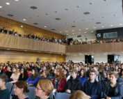 Ein großer Hörsaal der Hochschule Coburg ist voller aufmerksamer sitzender und stehender Studenten. Der Raum mit seiner Holzvertäfelung und seinem modernen Design verfügt über abgestufte Sitzbereiche und helles Licht, was eine inspirierende Lernumgebung schafft.