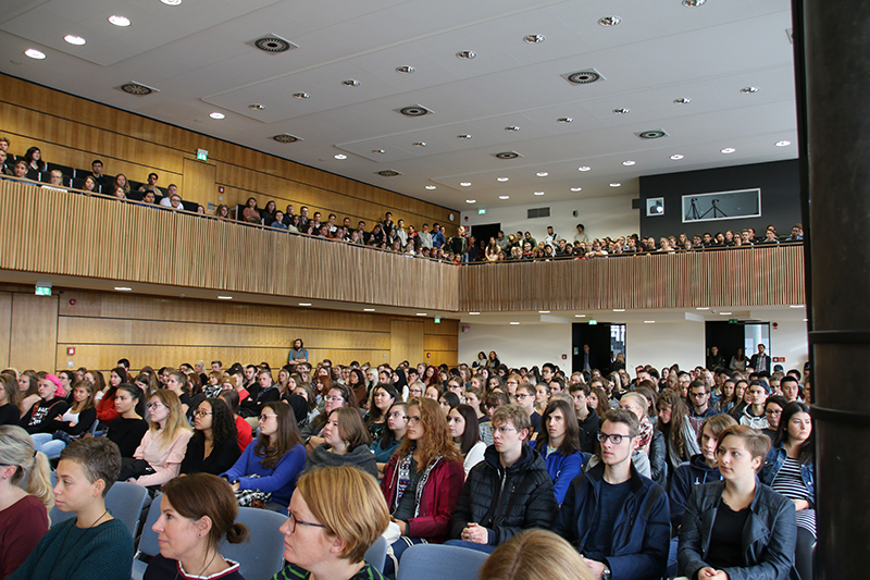 Ein großer Hörsaal der Hochschule Coburg ist voller aufmerksamer sitzender und stehender Studenten. Der Raum mit seiner Holzvertäfelung und seinem modernen Design verfügt über abgestufte Sitzbereiche und helles Licht, was eine inspirierende Lernumgebung schafft.