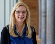 Eine Frau mit langen blonden Haaren und Brille lächelt, trägt ein blaues Hemd und einen schwarzen Pullover. Sie steht in einem Flur, im Hintergrund ist eine Holzvertäfelung zu sehen, die an die elegante Architektur der Hochschule Coburg erinnert.