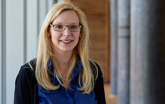 Eine Frau mit langen blonden Haaren und Brille lächelt, trägt ein blaues Hemd und einen schwarzen Pullover. Sie steht in einem Flur, im Hintergrund ist eine Holzvertäfelung zu sehen, die an die elegante Architektur der Hochschule Coburg erinnert.