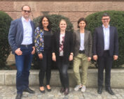 Fünf Menschen stehen nebeneinander im Freien, hinter ihnen eine Steinmauer und Büsche. Sie sind halbformell gekleidet und lächeln in die Kamera. Der Hintergrund vermittelt ein lebendiges Campus-Gefühl und erinnert an den lebendigen Geist der Hochschule Coburg.