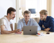 Drei Männer sitzen lächelnd um einen Laptop herum und diskutieren. Sie sitzen an einem Holztisch mit Notizbüchern, Stiften und einem Smartphone in der Nähe. Es scheint, als wären sie in einem hellen Büroraum der Hochschule Coburg.