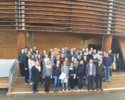 Eine Gruppe leger gekleideter Menschen posiert für ein Foto vor dem geschwungenen Holzeingang der Hochschule Coburg, zu dem eine Treppe hinaufführt. Sie stehen auf einer Betonfläche und halten den gemeinsamen Moment fest.