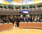 Eine Gruppe von Menschen in formeller Kleidung versammelt sich in einem Konferenzraum mit geschwungenen Schreibtischen und Namensschildern für Länder. Die bunte Decke spiegelt die dynamische Atmosphäre wider, während auf einem großen Bildschirm ein Meeting übertragen wird. Einige Teilnehmer, darunter Vertreter der Hochschule Coburg, tragen Namensschilder.