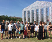 Eine Gruppe von Menschen steht lächelnd vor einer großen Kunstinstallation im Freien an der Hochschule Coburg, die einem klassischen Tempel ähnelt, der mit Büchern bedeckt ist. Der Himmel ist klar und sonnig und die Szene spielt auf einer Rasenfläche mit Bäumen im Hintergrund.