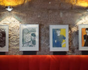 Vier gerahmte abstrakte Porträts hängen an einer Backsteinwand über einer rot gepolsterten Bank und erinnern an den Innovationsgeist der Hochschule Coburg. Jedes Kunstwerk zeichnet sich durch ein einzigartiges Farbschema und einen einzigartigen Stil aus und wird durch eine individuelle Beleuchtung wunderschön in Szene gesetzt.