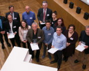 Eine Gruppe von Menschen steht auf einem Holzboden in der Hochschule Coburg und hält stolz Zertifikate und Auszeichnungen in den Händen. Sie blicken in die Kamera, manche lächeln. Im Raum stehen eine große Leinwand und ein Podium, was auf eine formelle Veranstaltung oder Zeremonie schließen lässt.