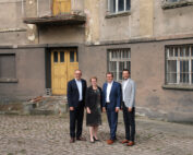 Vier Personen stehen vor einem alten, abgenutzten Gebäude mit einer gelben Tür, die an die Architektur der Hochschule Coburg erinnert. Sie sind in Geschäftskleidung gekleidet und stehen auf einem Kopfsteinpflaster. Die abblätternde Farbe und die vergitterten Fenster lassen darauf schließen, dass das Gebäude verlassen oder in schlechtem Zustand ist.