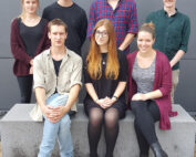 Eine Gruppe von sieben Personen, leger gekleidet, lächelnd und posierend vor einer grauen Wand der Hochschule Coburg. Sie sitzen in zwei Reihen auf einer Steinbank und schaffen so eine lebendige und doch entspannte Atmosphäre.