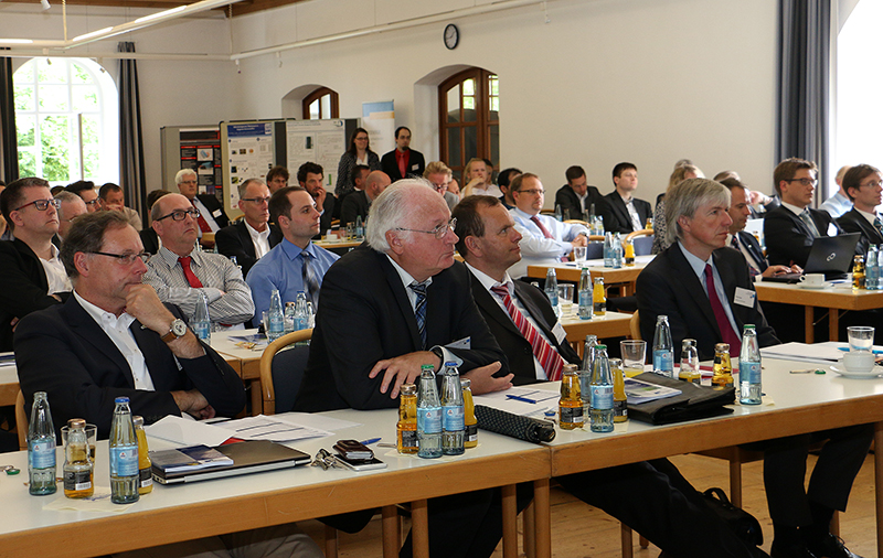 Eine Gruppe von Männern und Frauen sitzt aufmerksam in einem Konferenzraum der Hochschule Coburg und hört sich eine Präsentation an. Der Raum ist mit langen Tischen gefüllt, an denen Wasserflaschen und verstreute Papiere stehen. Die Teilnehmer sind in formelle Kleidung gekleidet, was das professionelle Ambiente der Hochschule widerspiegelt.